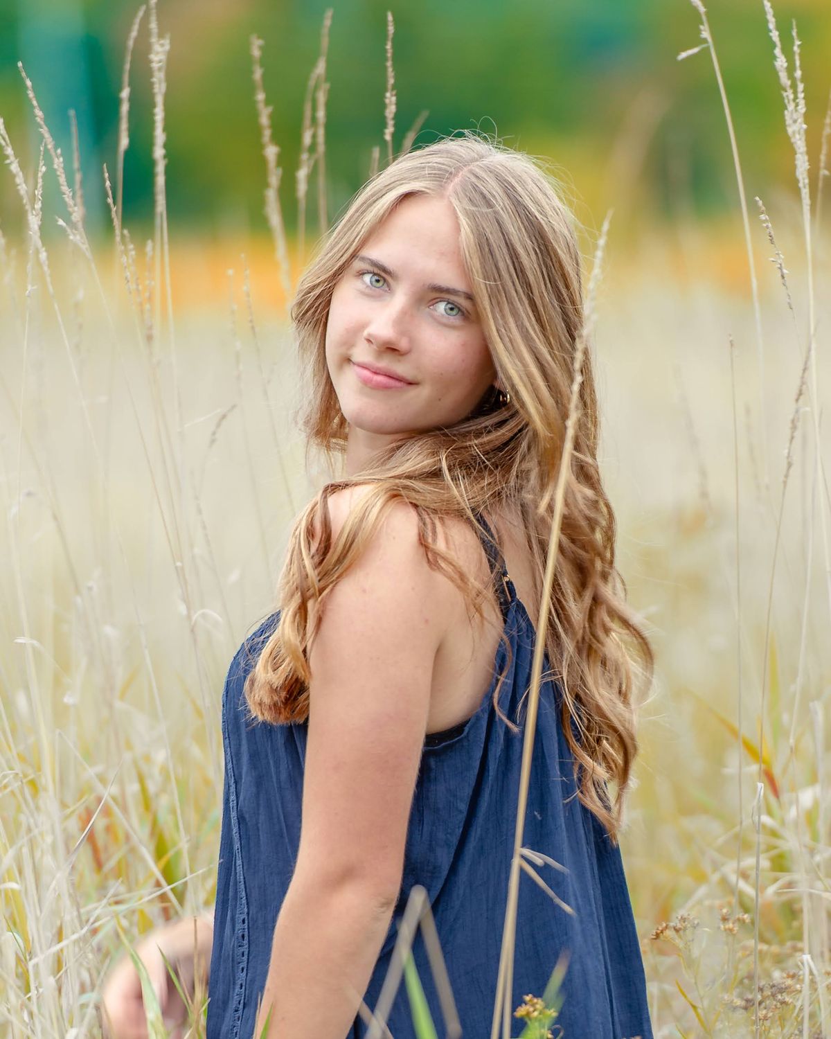 A teenage girl in a blue dress standing in tall golden grass, looking back over her shoulder