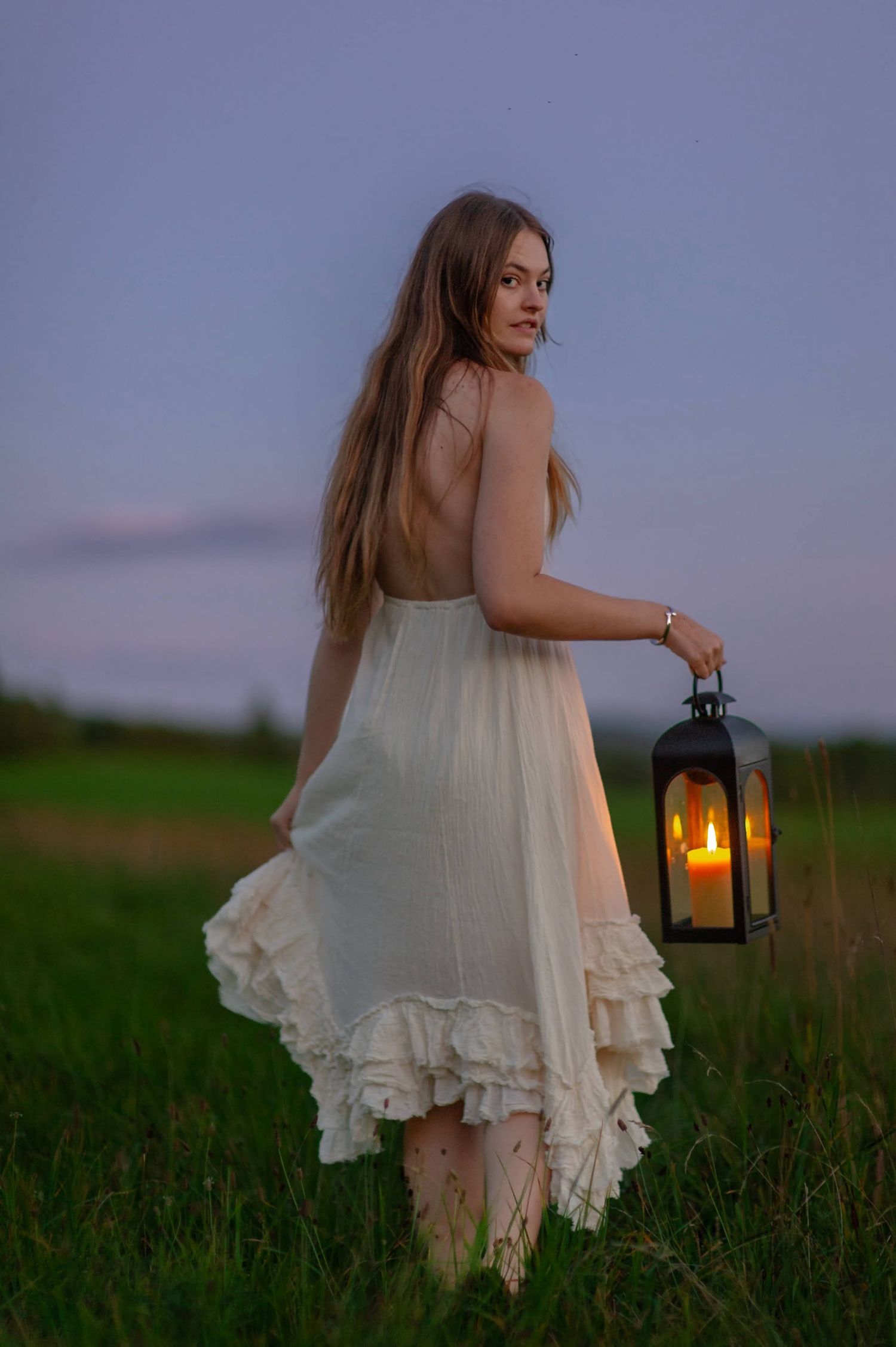 A young woman in a pale dress at dusk, carrying a lit lantern through tall grass