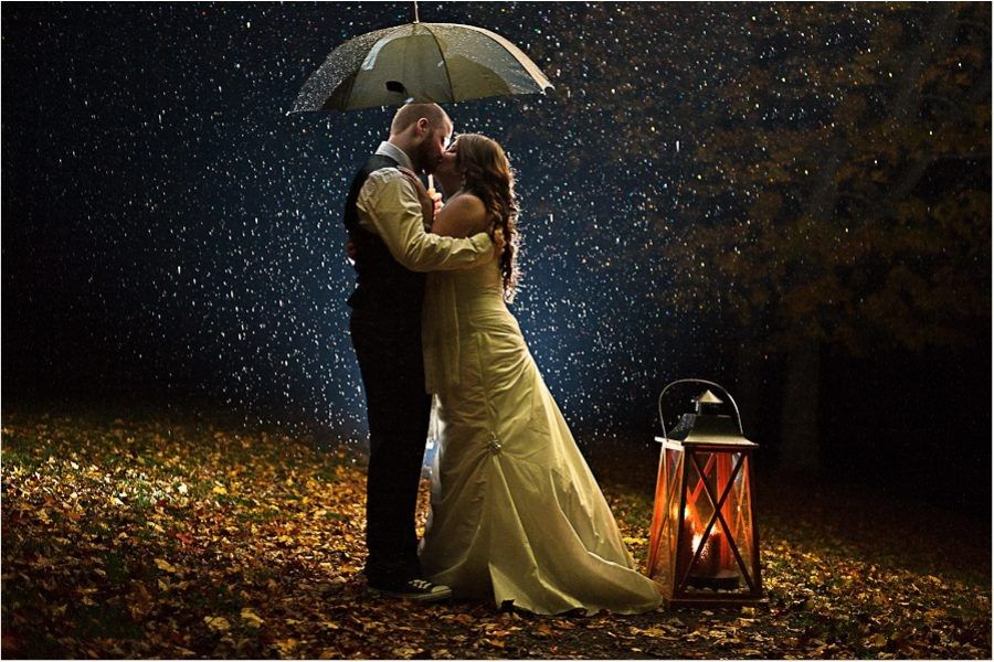A couple under an umbrella in heavy rain at night, lit from behind so the raindrops glow, a lantern beside them